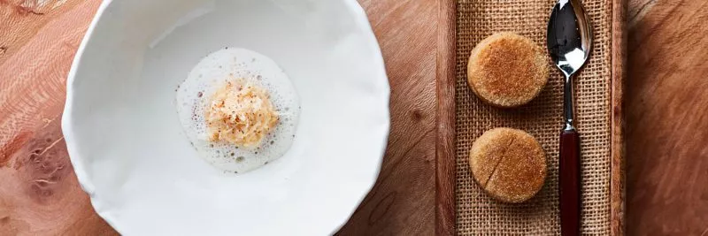 Top down view of a table at Restaurant St. Barts, a white ceramic shallow bowl with a delicate dish with foam, two small rolls of bread ad a spoon on the side.